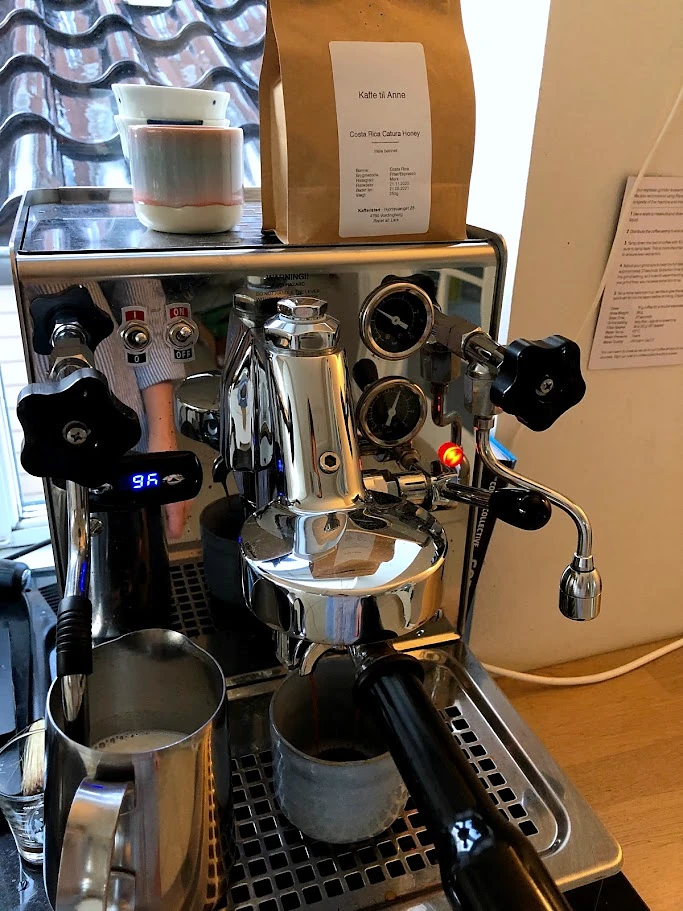 A chrome espresso machine with two gauges, a digital thermometer, cups and a coffee bag