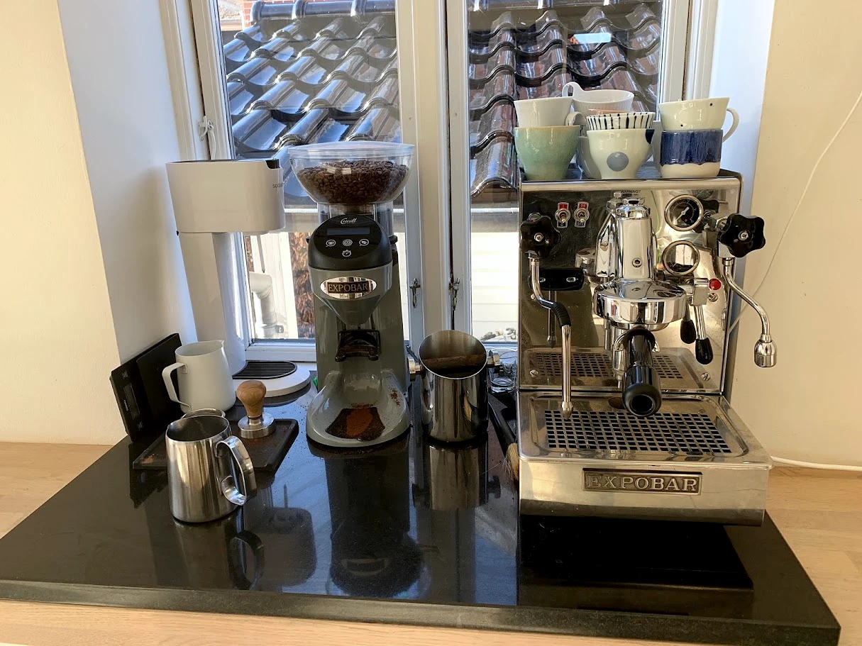 An Expobar espresso machine and hopper grinder with cups and milk pitchers on a home coffee bar
