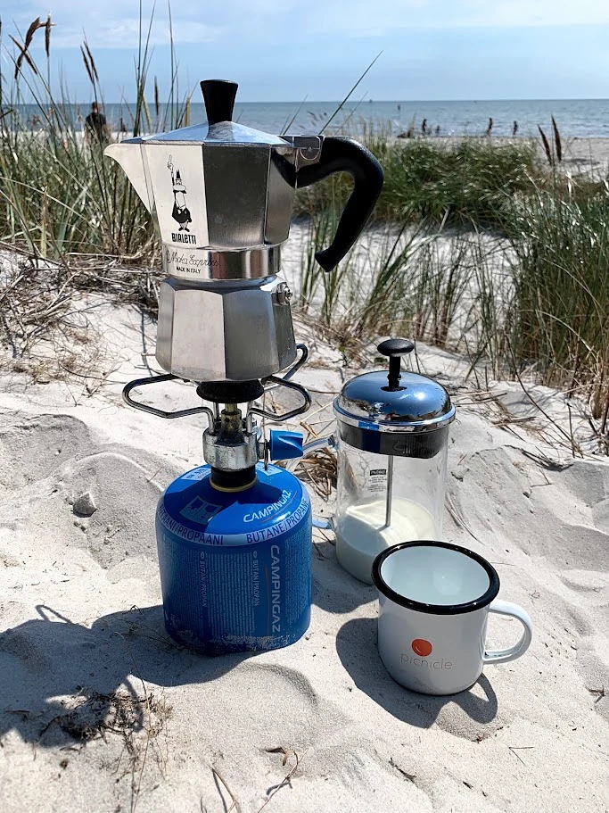A moka pot on a portable stove with a mug and camping coffee equipment beside the beach