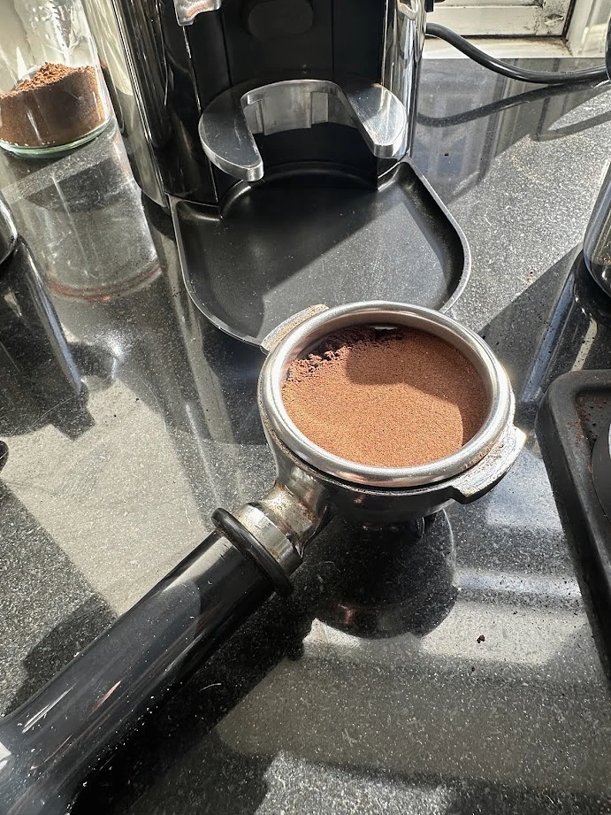 A portafilter basket filled with ground coffee in front of a grinder